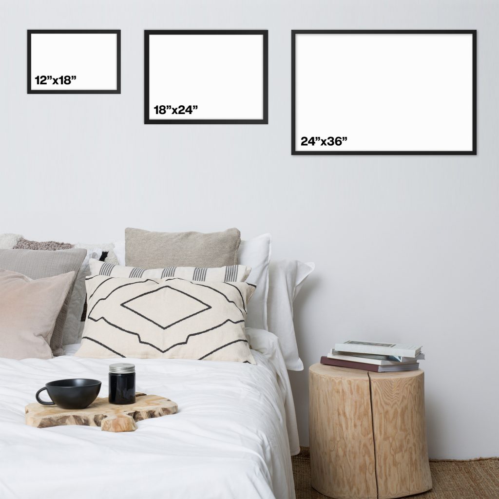 Three framed blank canvases of different landscape sizes above a neatly made bed with a round wooden side table.